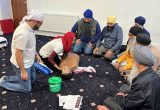 Rohit Sagoo (left) founder of the British Sikh Nurses group, teaching CPR to Sikh men in a local gurdwara. There are six men gathered around a CPR dummy.